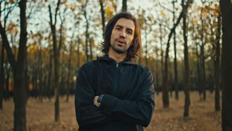 Confident-serious-man-with-medium-length-hair-and-a-brunette-beard-holds-his-arms-crossed-on-his-chest-in-a-black-sports-uniform-stands-in-the-autumn-forest