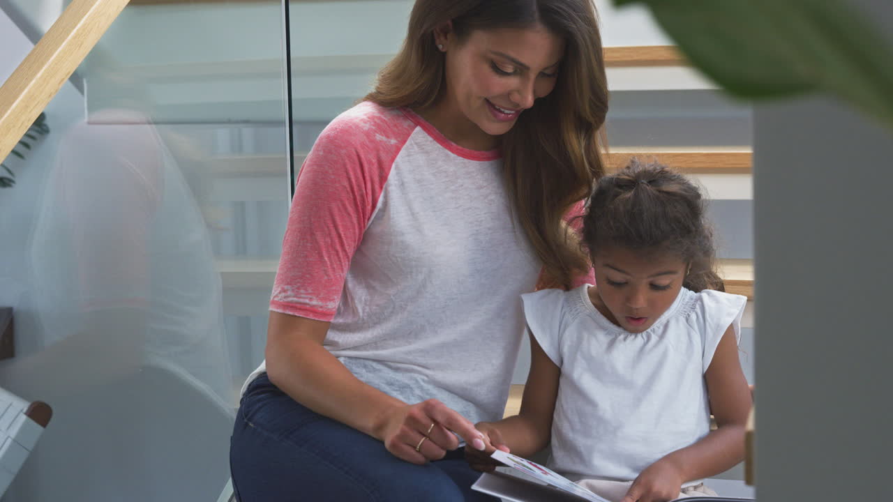 madre y hija hispanas sentadas en la escalera en una casa moderna leyendo un libro juntas