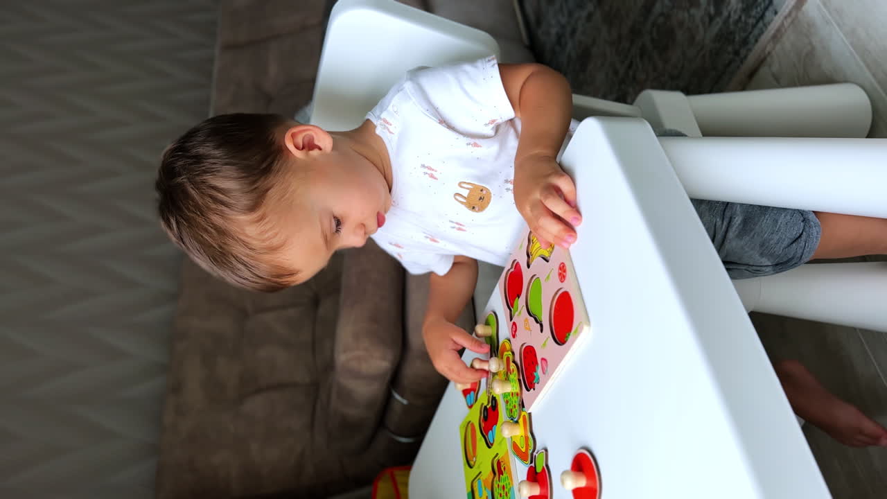 Adorable Caucasian boy sitting at the desk in his room. Baby playing peacefully with puzzles of fruit. Vertical screen.