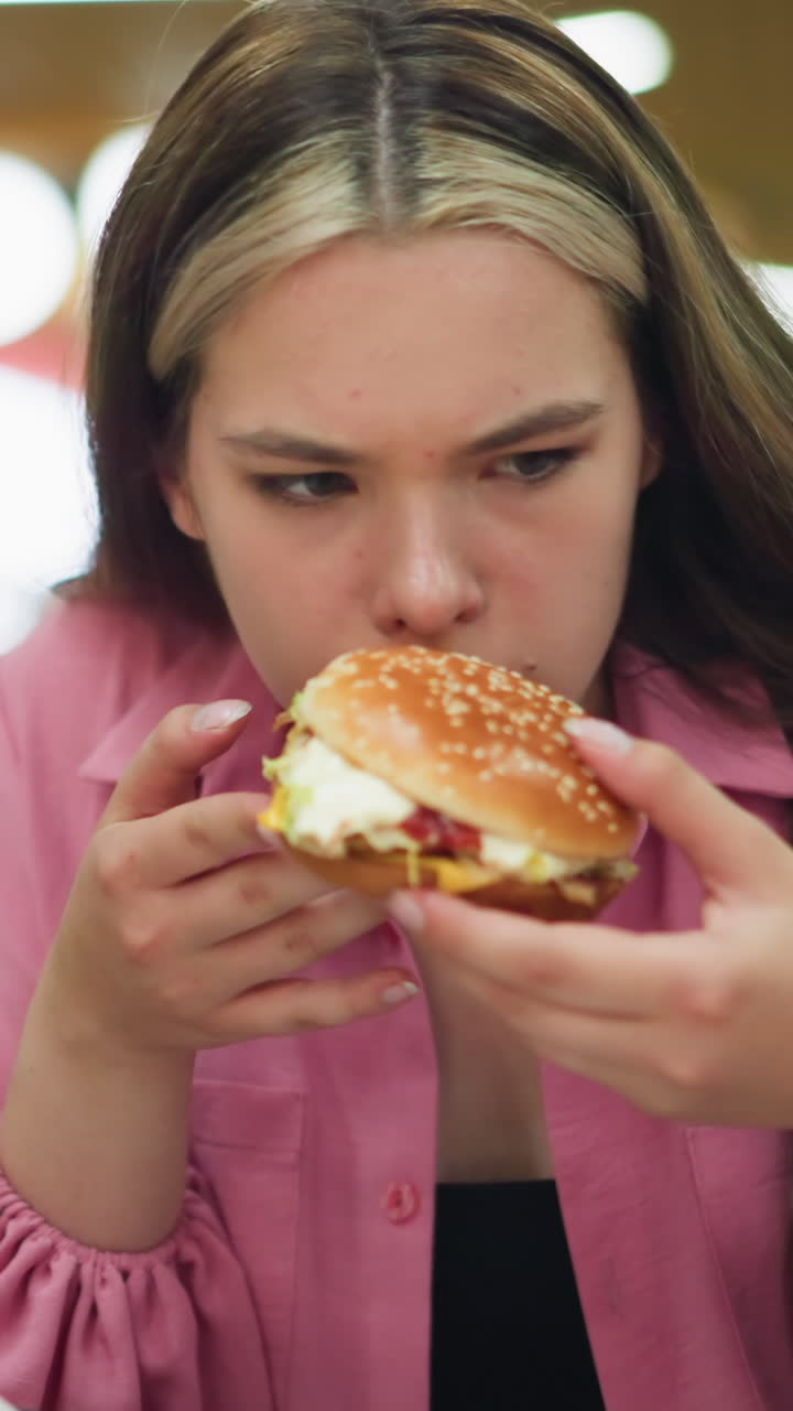 dama de vestido rosa huele hamburguesa con disgusto, mostrando asco después de dejarlo caer, ella alcanza una servilleta para limpiar su mano mientras la gente en el fondo continúa comiendo en una vista borrosa