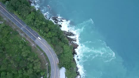 Top-aerial-view-following-road-with-trees-alongside-rocky-shoreline-in-Thailand