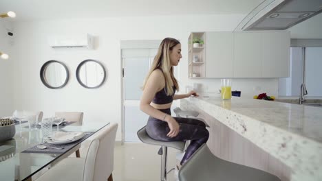 young blonde woman in sportswear sitting in a luxurious kitchen to have a juice for breakfast