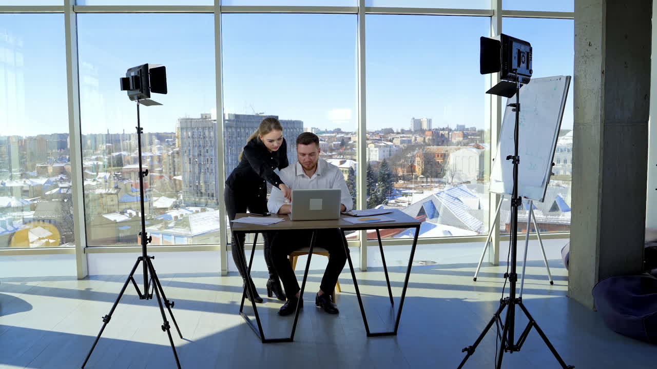 Business colleagues working together in office. Businessman is sitting at table and working on a laptop. Young woman helps a man in business. City landscape through the window.