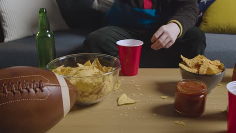 Persona-En-Casa-Viendo-Un-Partido-De-Fútbol-Americano-En-La-Televisión-Con-Bebidas,-Bocadillos-Y-Pelota