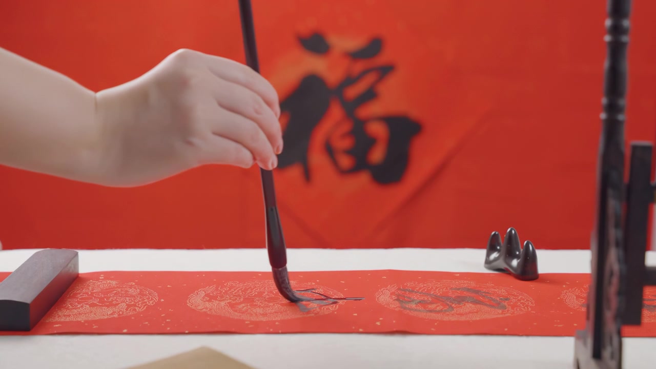 Mão de artista praticando caligrafia chinesa tradicional usando tinta dourada em papel carmesim, pinceladas habilidosas criam caracteres elegantes que transmitem sabedoria em uma mesa de madeira.