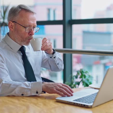 Senior man with a cup working at home on laptop. Freelancer using computer from home-office. Mature businessman working near the window.