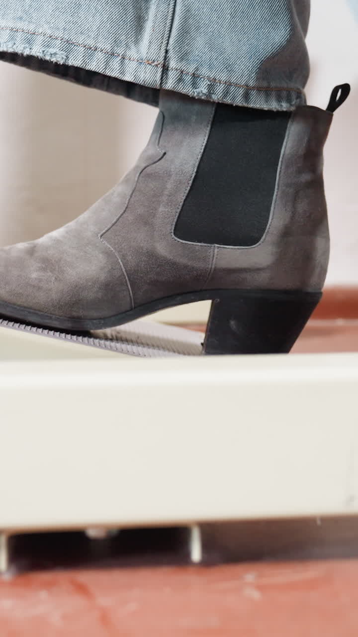 primer plano de bota Chelsea pisando el pedal, bota gris de ante con paneles laterales elásticos en contacto con el pedal texturizado sobre suelo de terracota, bucle de movimiento dinámico, detalle de moda moderna, ambiente de estudio energético, tacón