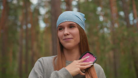 young woman with blue bandana smiles while brushing her hair in serene forest setting, radiating warmth and natural beauty, surrounded by lush trees, she enjoys a peaceful moment in nature