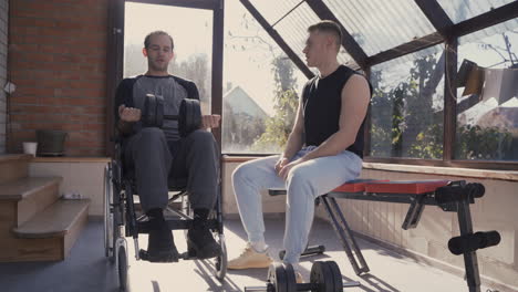 Disabled-Man-In-Wheelchair-Working-Out-With-Dumbbell-At-Home-While-His-Personal-Trainer-Motivating-Him