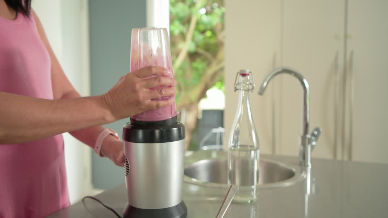 Kvinne heller frukt i kopp på benken, trykker for å blande rosa smoothie, setter på lokk for å drikke.