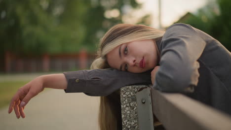 woman in grey clothing resting her head on arm outdoor, lost in thought with a peaceful expression, the background is blurred, highlighting her thoughtful state