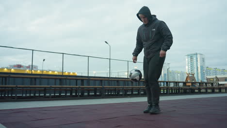 man skillfully juggling soccer ball on outdoor sport arena with chain-link fence and benches in background, urban buildings and playground visible in distance