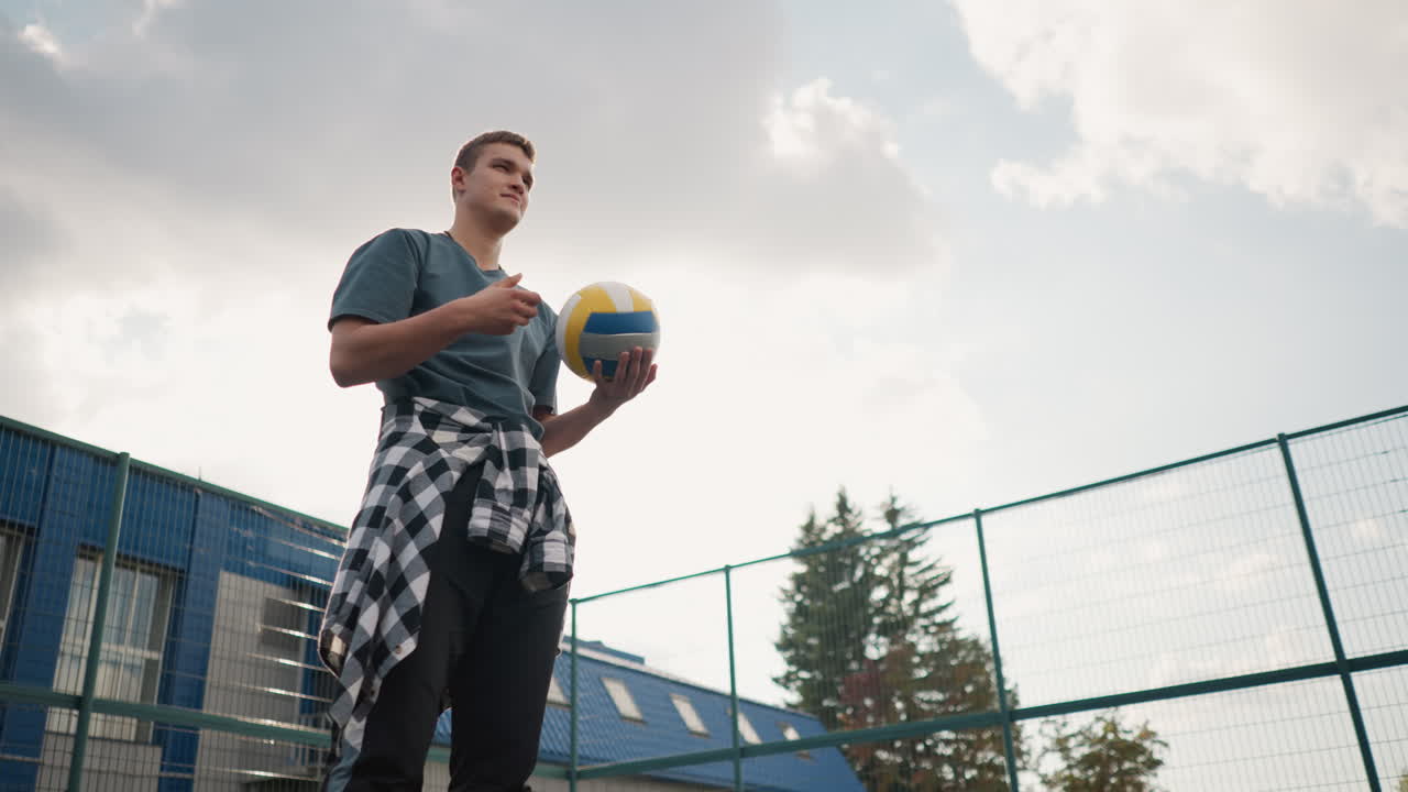 joven con camisa a cuadros alrededor de la cintura gira la pelota de voleibol en la mano, con árboles y cancha deportiva al aire libre en el fondo, listo para la acción
