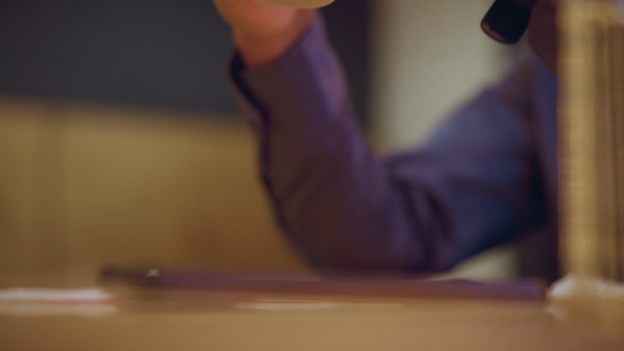 Blurred close-up of person s arm in purple shirt lifting hand while smartphone rests on wooden table in foreground, soft ambient lighting creates quiet indoor setting conveying pause, stillness, or anticipation