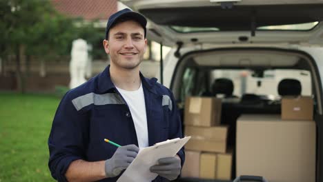 feliz sonriente trabajador de servicio de entrega en uniforme, gorra y guantes hace notas en los documentos y de pie en la calle cerca de la