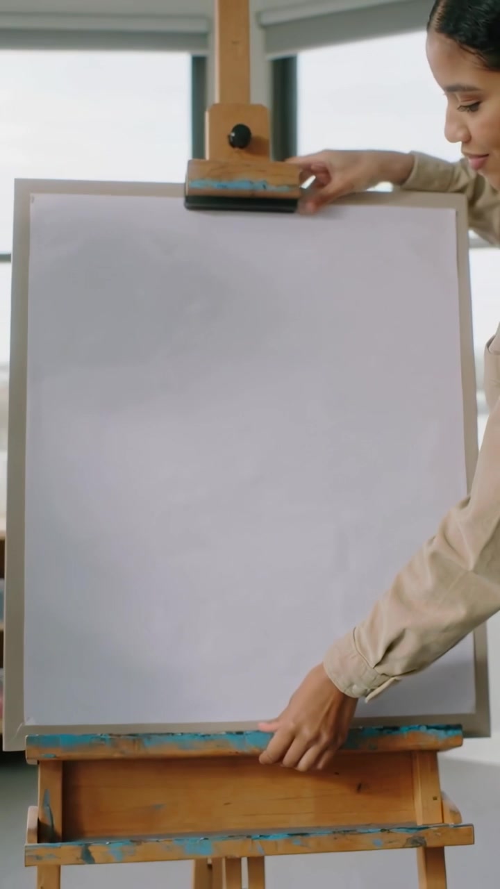 Video verticale: Donna con top color cuoio che si allunga, usa una pinza per allineare una tela su un cavalletto in studio per dipingere
