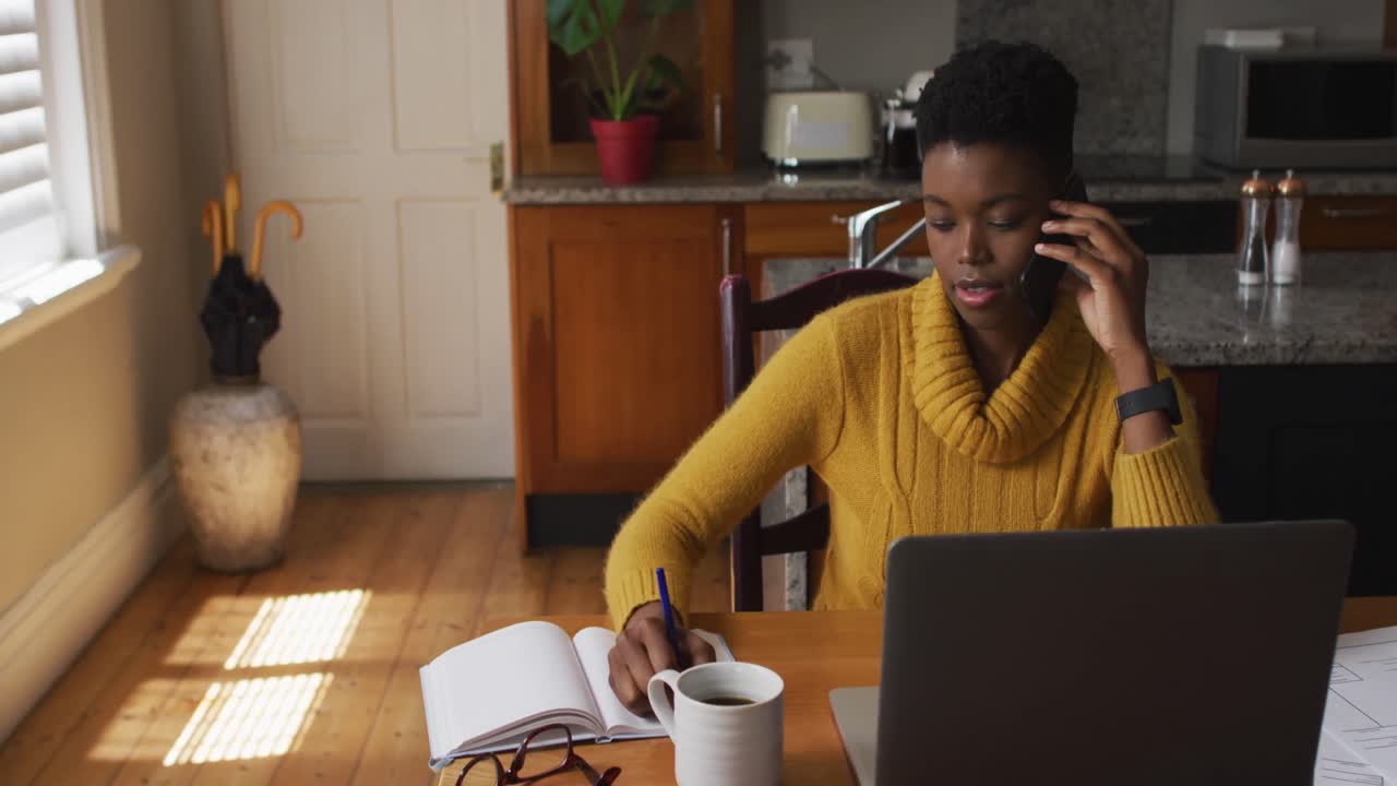 mujer afroamericana hablando en un teléfono inteligente y tomando notas mientras trabaja desde casa