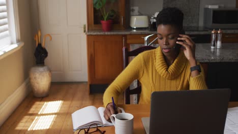 Afroamerikanische-Frau,-Die-Auf-Dem-Smartphone-Spricht-Und-Sich-Notizen-Macht,-Während-Sie-Von-Zu-Hause-Aus-Arbeitet