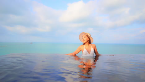 Woman-Model-in-White-Swimming-Suit-Leaning-Arms-on-the-Edge-of-Infinity-Pool-on-Turquoise-Seascape-Background-in-Bali-on-a-Sunny-Day-Slow-Motion