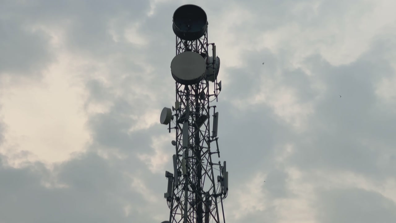 A tight tilt-up shot of a telecom tower showing antennas and satellite dishes, with a bird perched on one of the disks against a cloudy sky.