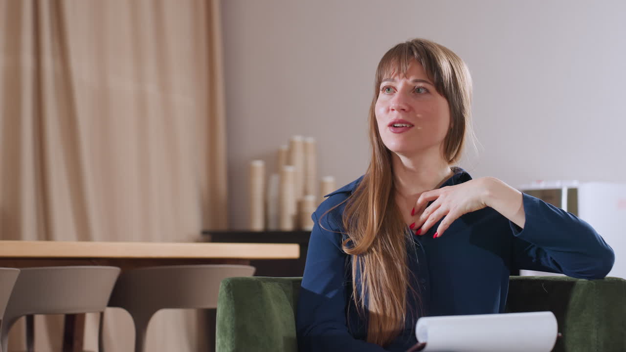 Specialist seated in green chair explaining from notes while speaking during therapy session, modern room with wooden table chairs, window showing dusk sky and beige curtains in indoor environment