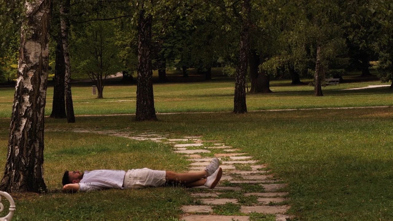 Young man sits on the grass before lying down to relax under trees in a green park, enjoying a warm summer day and quiet leisure time outdoors.