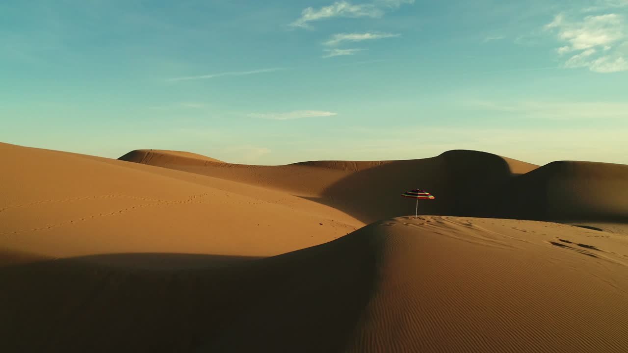 Aerial fly by of colorful beach umbrella isolated in desert sand dunes at sunset
