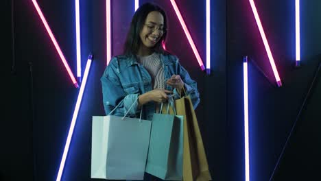 young girl in blue denim jacket with shopping bags on neon lamps background.