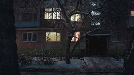 office building exterior illuminated by glowing light streaming through windows surrounded by snow-covered branches of trees, under dim evening sky