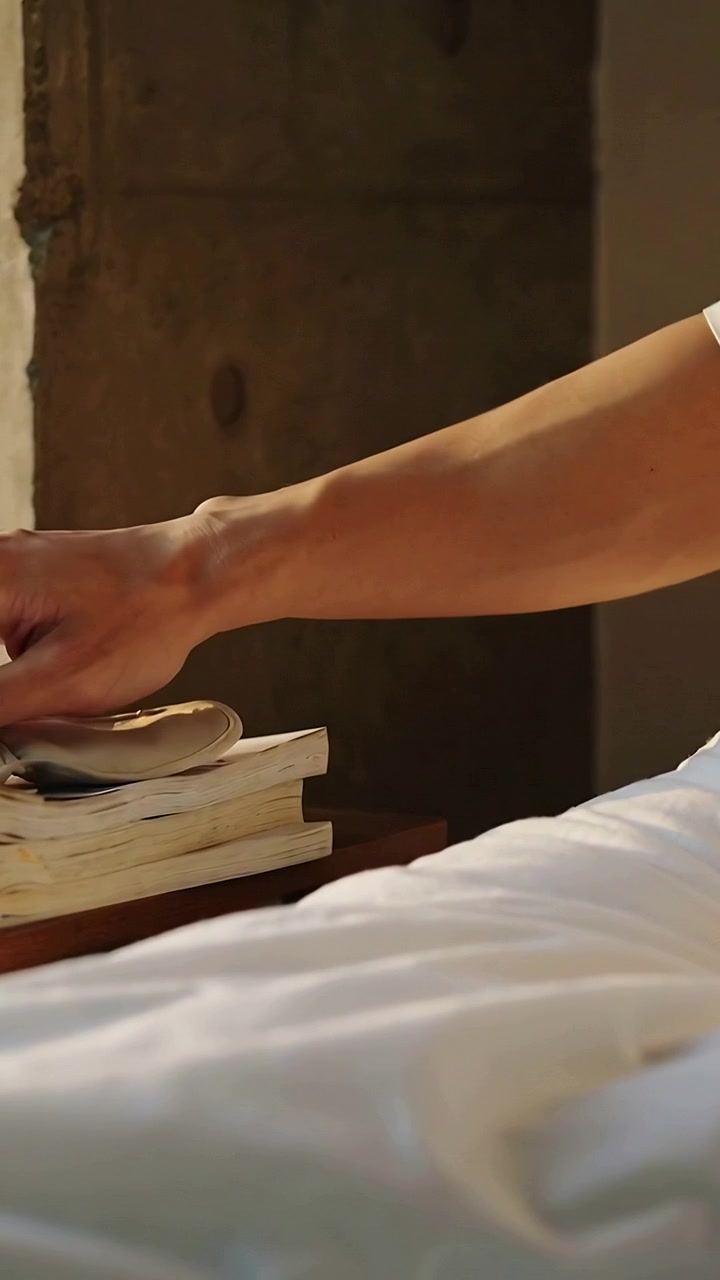 Video verticale: Mano destra con maglietta bianca che si allunga sollevando un dispositivo argentato dai libri sul comodino.