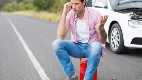 hombre caucásico sentado en un bote hablando en un teléfono inteligente por un coche averiado en una carretera rural