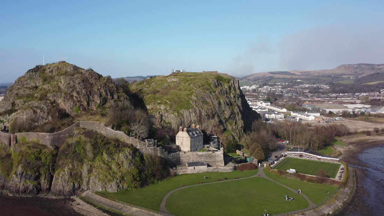 Aerial drone shot captures Dumbarton Castle in Scotland built on steep volcanic rock above river, showing fortified walls, historic buildings, green lawn, and distant landscape under daylight