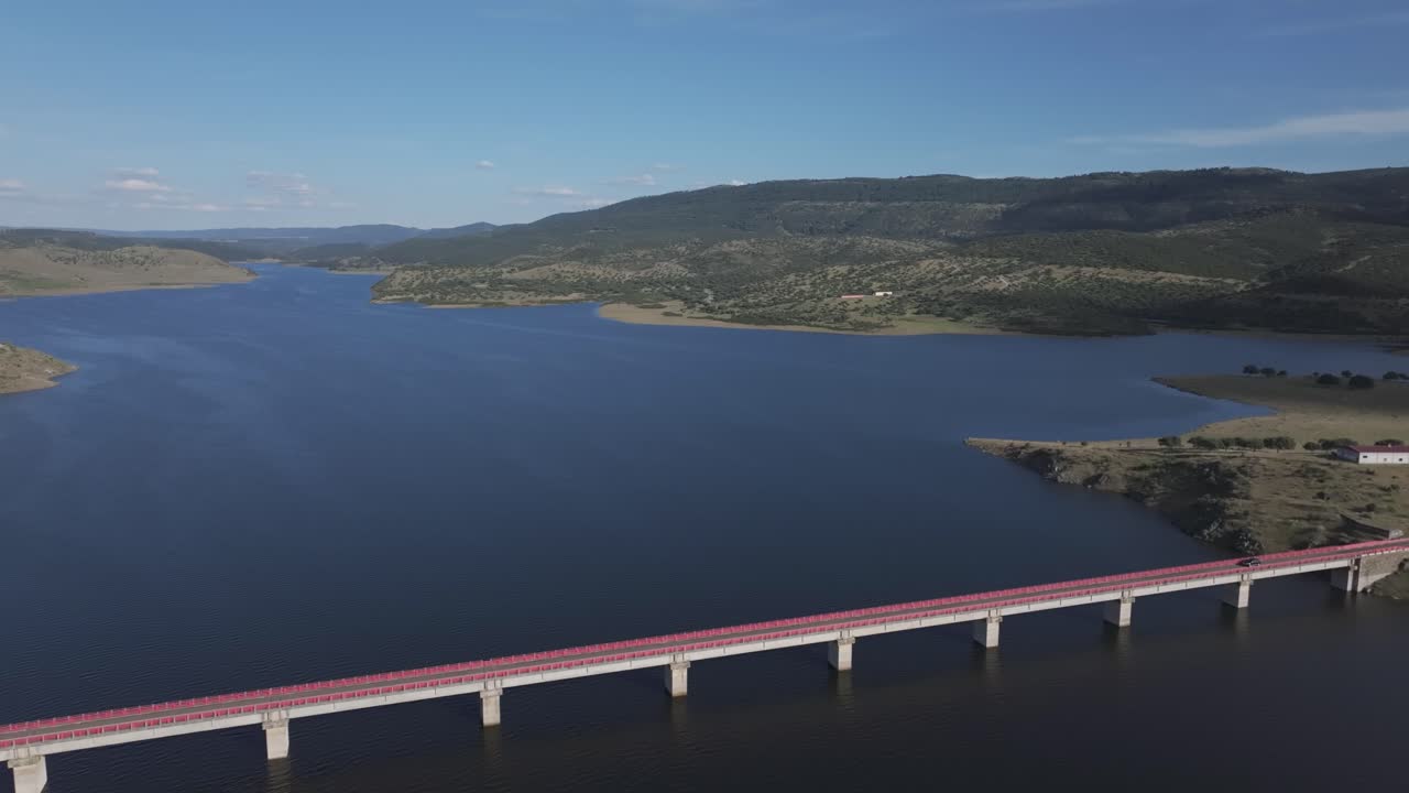 Drone view of the Bridge of La Mesta spanning the Cíjara reservoir in Extremadura, Spain. Its red railings and ongoing vehicle traffic contrast with the still water and natural surroundings