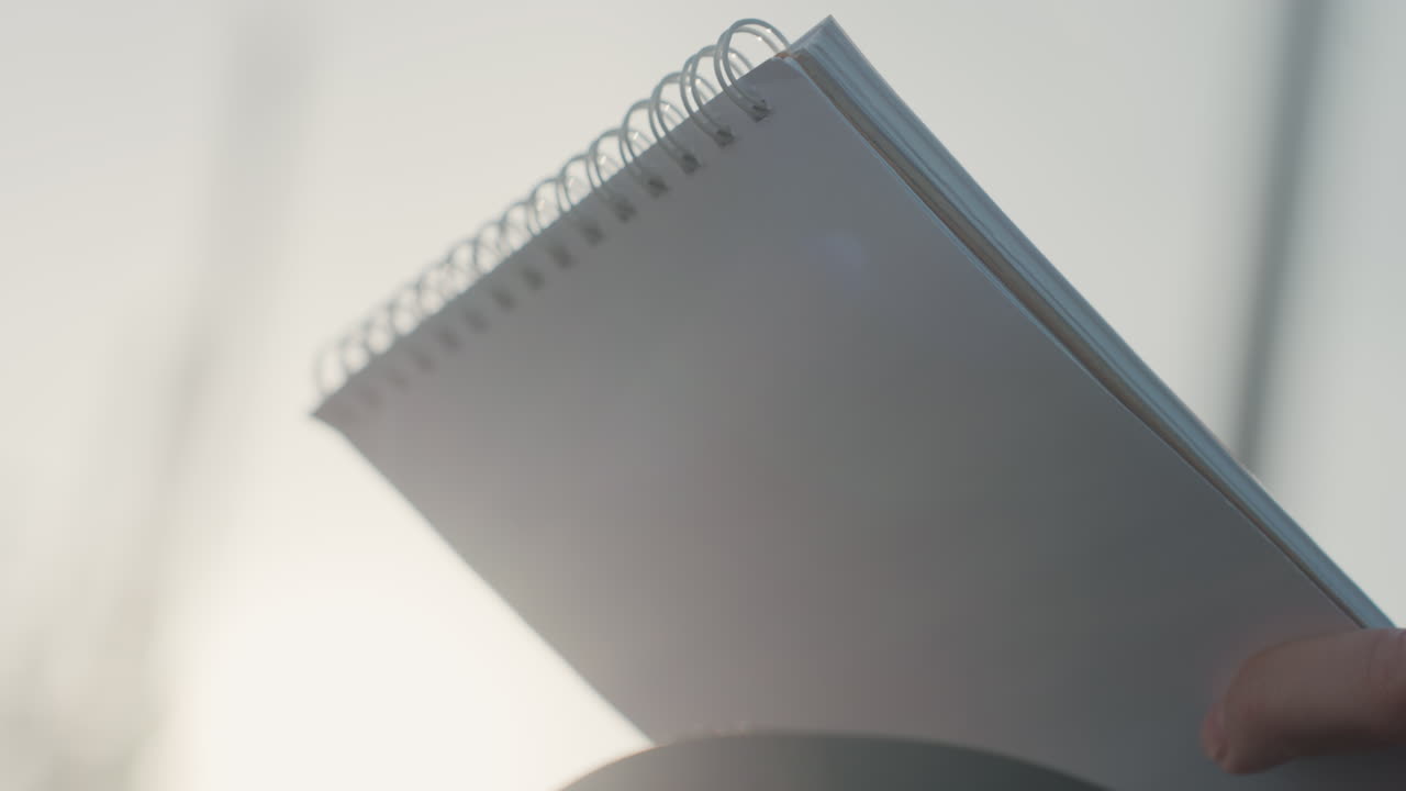 Primo piano di una mano bianca che tiene un quaderno con rilegatura a spirale e pagine vuote, illuminato dal controluce di una luce solare soffusa, profondità di campo ridotta, composizione equilibrata, con un senso di tranquilla attesa e atmosfera minimalista, in attesa di uno schizzo