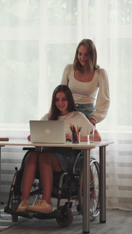 blonde woman helps girl partner with disability in wheelchair get to table with laptop. happy ladies girlfriends hug expressing love and support at home