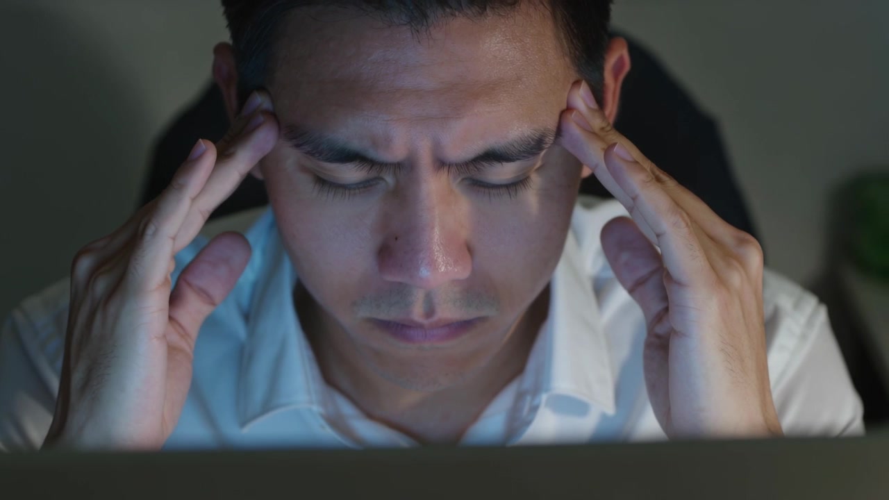 Reagendo al bagliore del laptop, uomo asiatico in camicia bianca che si massaggia le tempie cercando sollievo alla scrivania dell'ufficio