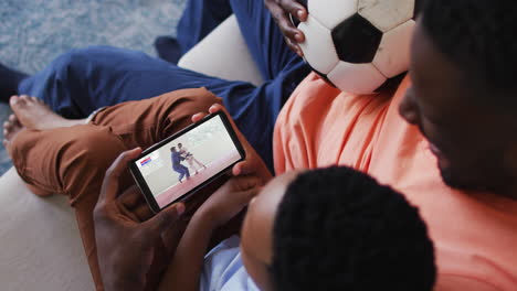 composite of father and son at home watching judo match on smartphone