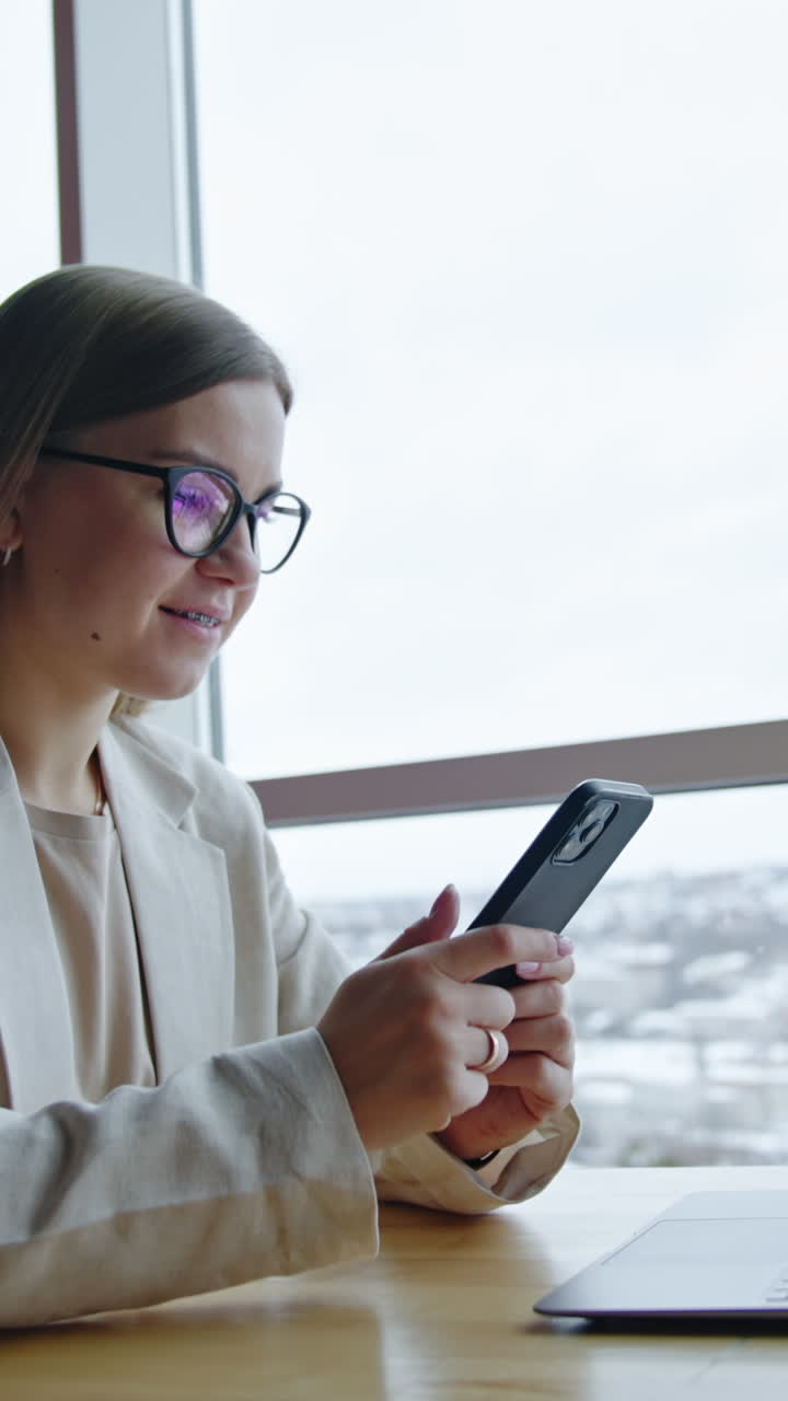 Young woman sitting at desk near the window using gadgets. Working lady uses smartphone for business communication. Vertical video