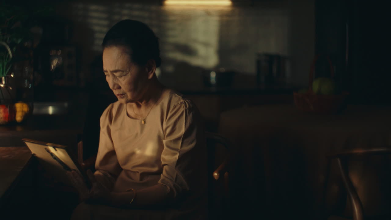 Older woman sitting in dimly lit room, looking at a photo with deep reflection