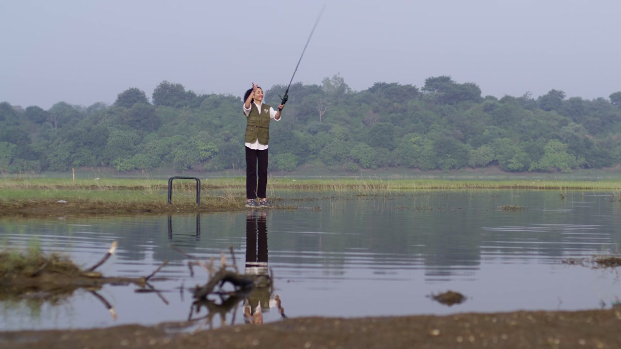 A South Asian woman enjoys fishing at sunrise near tranquil waters beneath a clear pastel morning sky