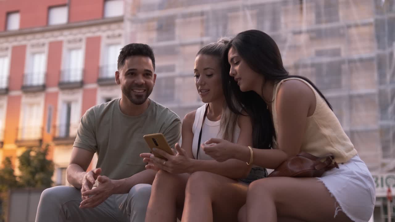 Friends talking while using smartphone together on sidewalk in city during sunset