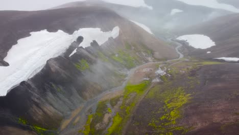 aerial drone footage flying over dark brown and green mossy iceland volcanic terrain landscape with snow on steep, big mountains and a narrow river flowing between them during a cloudy and misty day.