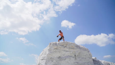 shirtless athlete lifting kettlebell. healthy sportsman is swinging kettlebell with two hands on the top of white hill. view from below on bodybuilder under blue sky.