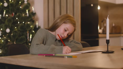 chica con suéter verde escribiendo una carta y pensando en deseos sentada en una mesa en una habitación decorada con un árbol de navidad 1