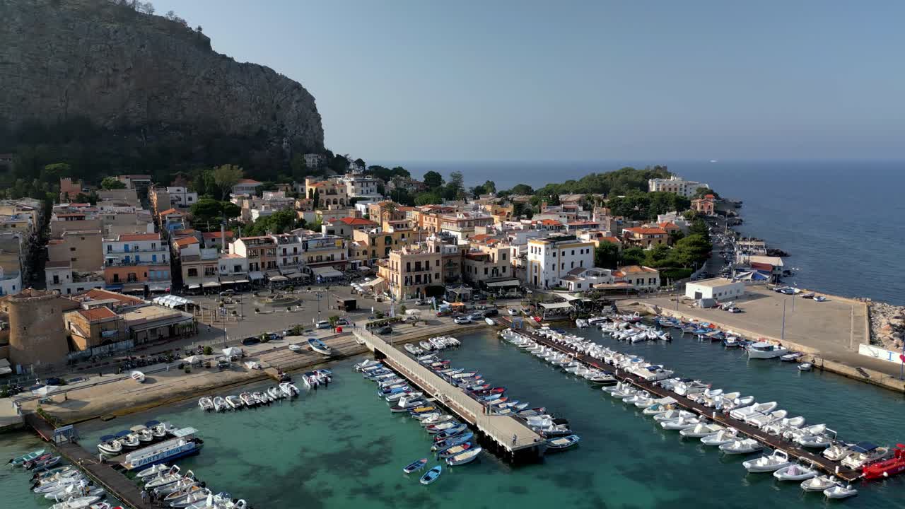 Ripresa drone ad alta angolazione che cattura la costa di Terrasini, il porto turistico e il limpido Mar Mediterraneo. Filmato drone che sorvola Terrasini evidenziando il contrasto tra l'acqua turchese bassa e il blu profondo.