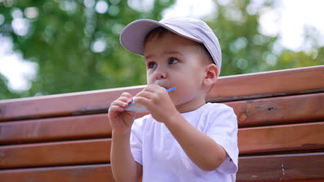 adorable kid wearing a cap holding a doy pack in his mouth. baby boy having a snack on the bench outdoors. low angle view.
