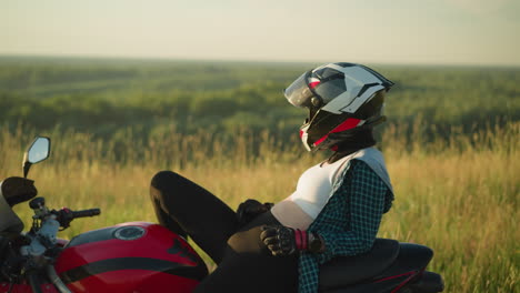 a young woman wearing a helmet rests on a red power bike in a field, adjusting her position slightly forward, her white crop top is visible, while the wind gently moves the surrounding grass