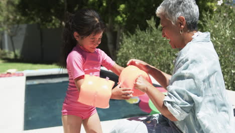 Una-Chica-Birracial-Con-Traje-De-Baño-Rosa-Vierte-Agua-De-Una-Jarra,-Asistida-Por-Su-Abuela