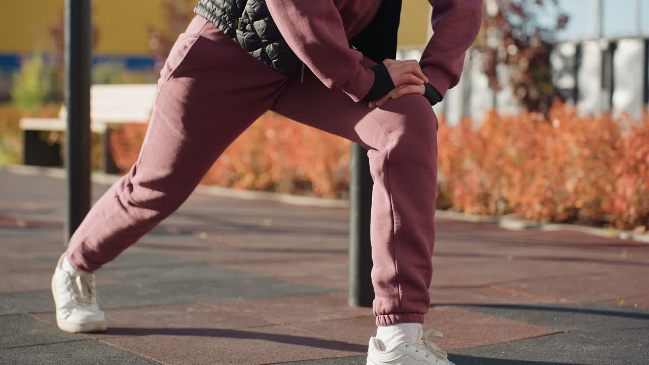 Exercise specialist in hoodie and vest performs forward pistol squat with hands braced on knee under bright sun casting long shadows across wide urban park tiles with blooming shrubs