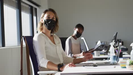 two diverse female colleagues wearing face mask, working at desk in office